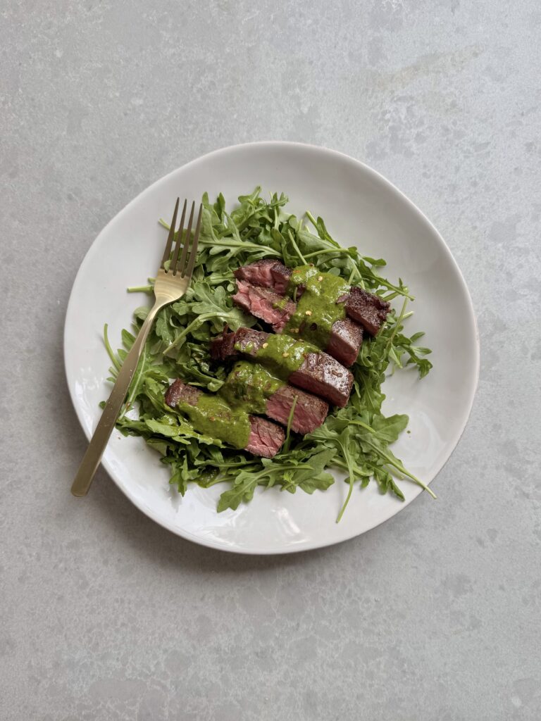 Sliced skirt steak topped with fresh chimichurri sauce on a cutting board – a high-protein dinner recipe by megannashhealth.