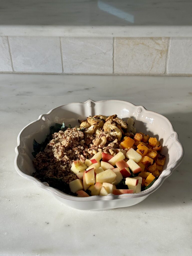 Harvest kale salad with roasted butternut squash, cauliflower, quinoa, walnuts, apple, and salmon drizzled with creamy tahini dressing on a white plate