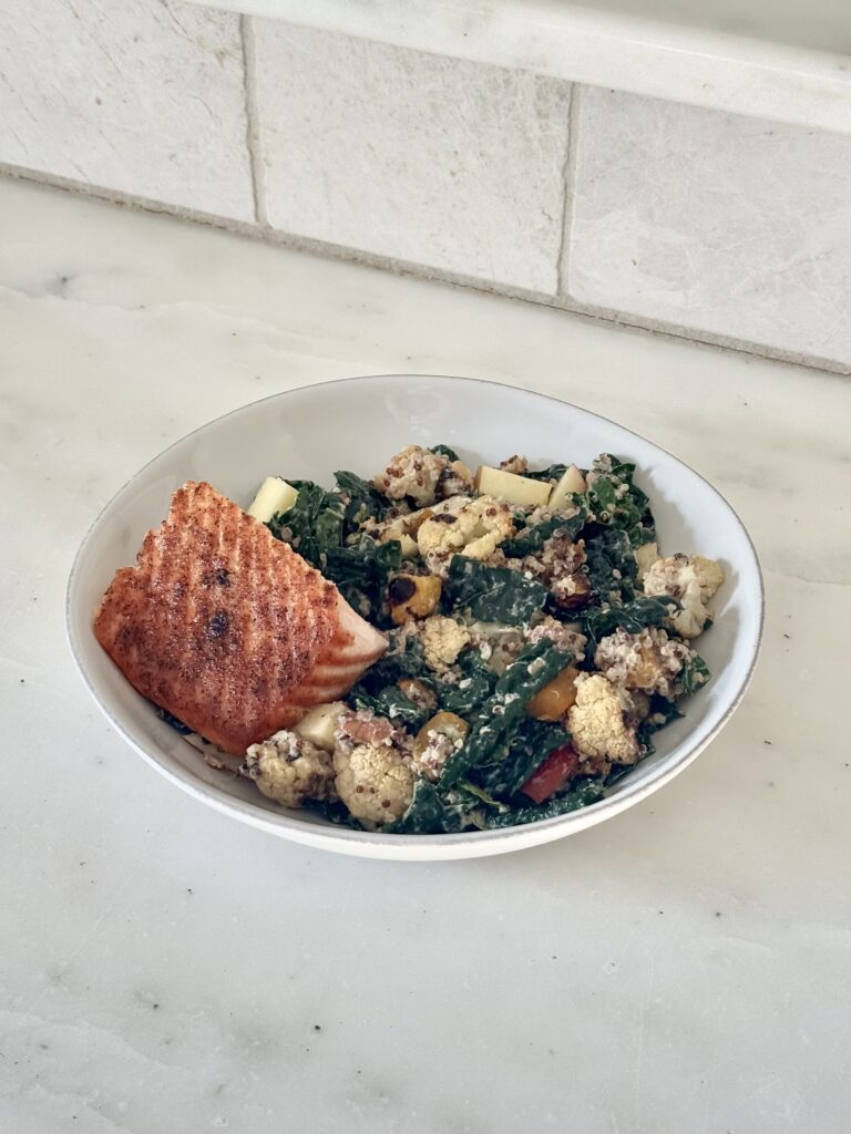 Harvest kale salad in a bowl topped with roasted vegetables, quinoa, apple, walnuts, and salmon with tahini dressing drizzled on top