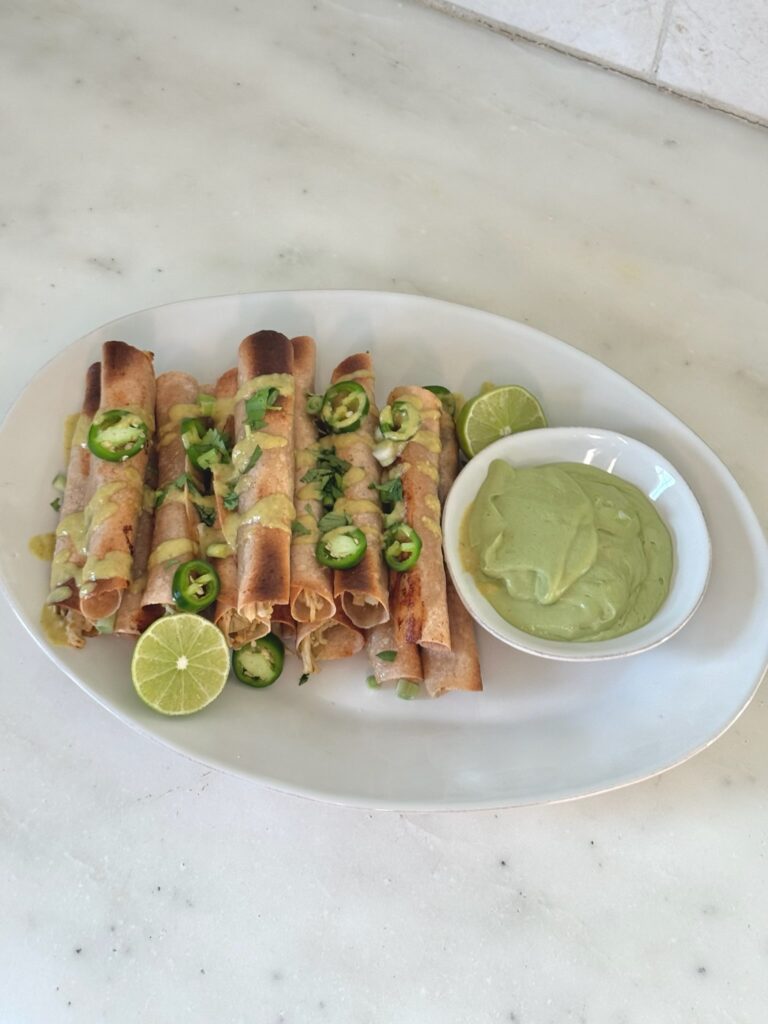 Plate of baked chicken taquitos topped with chopped jalapeños, green onion, cilantro, and creamy jalapeño sauce